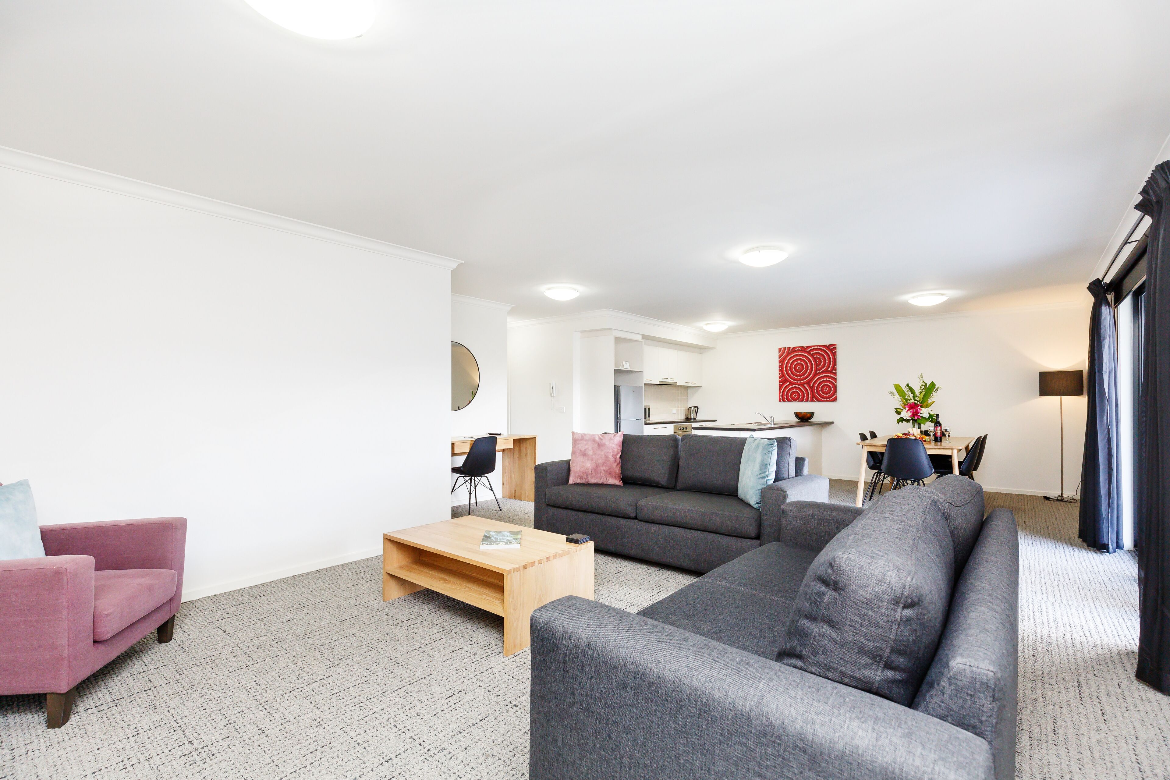 Een moderne woonkamer met een grijze bank, een roze fauteuil en een houten salontafel. Achtergrond is een keuken met een eettafel en stoelen.