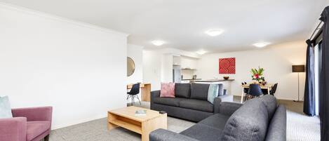 A modern living room with a grey sofa, a pink armchair, and a wooden coffee table. There is a kitchen area in the background with a dining table and chairs.