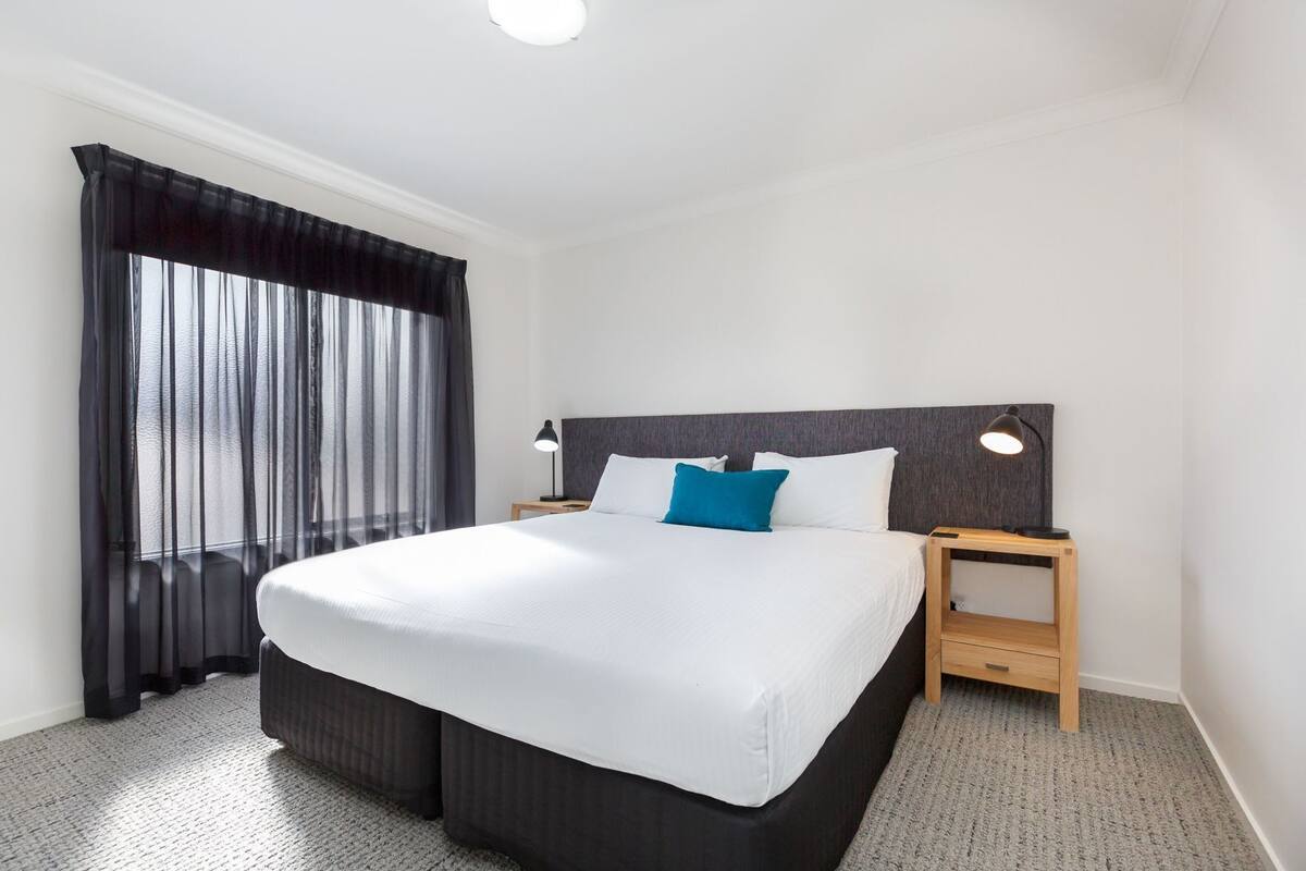 Una habitación de hotel con una cama grande, dos mesitas de noche, una lámpara de noche, una ventana con cortinas translúcidas y una luz en el techo.