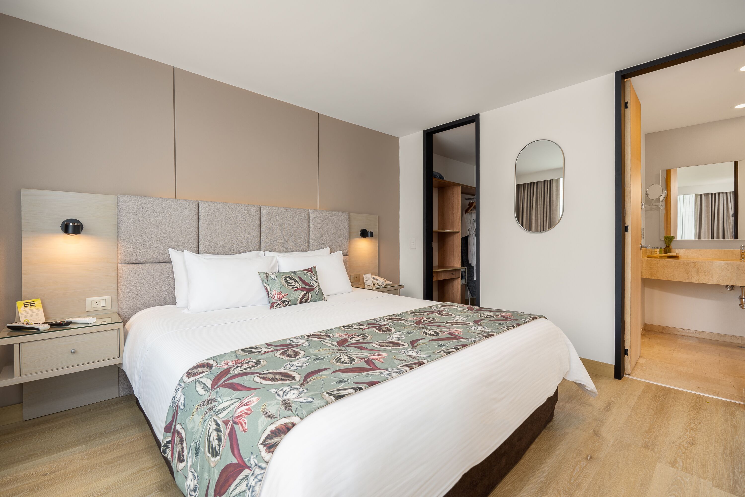 A modern hotel room with a large bed, bedside tables, a wardrobe, and a bathroom with a mirror and sink.