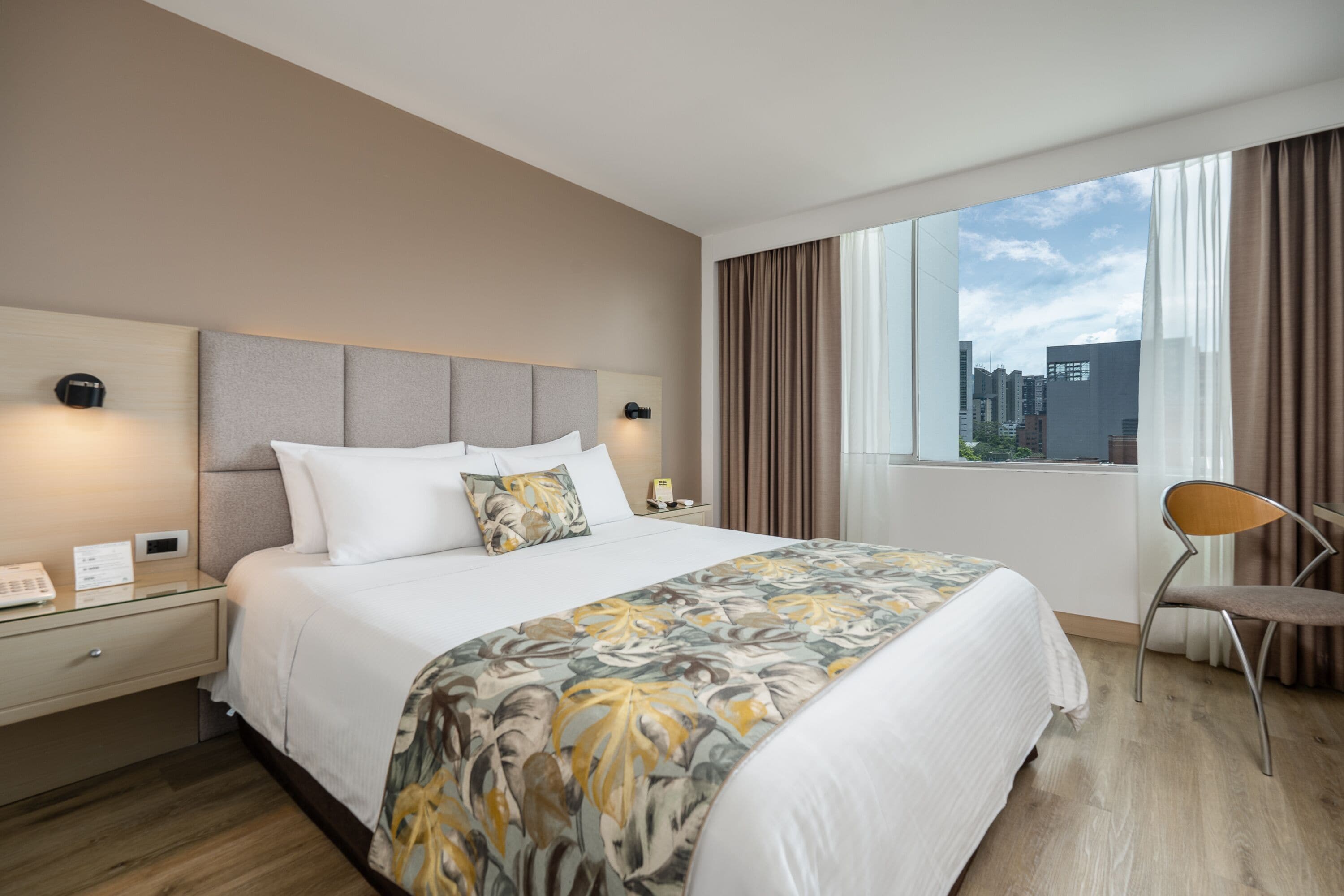A hotel room with a large bed, a nightstand, a chair, and a window with a city view.