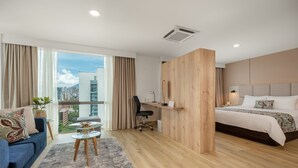 A modern hotel room with a large bed, a desk, a blue sofa, and a view of the city. - Novelty Suites Hotel - Oviedo Mall (Medellín)