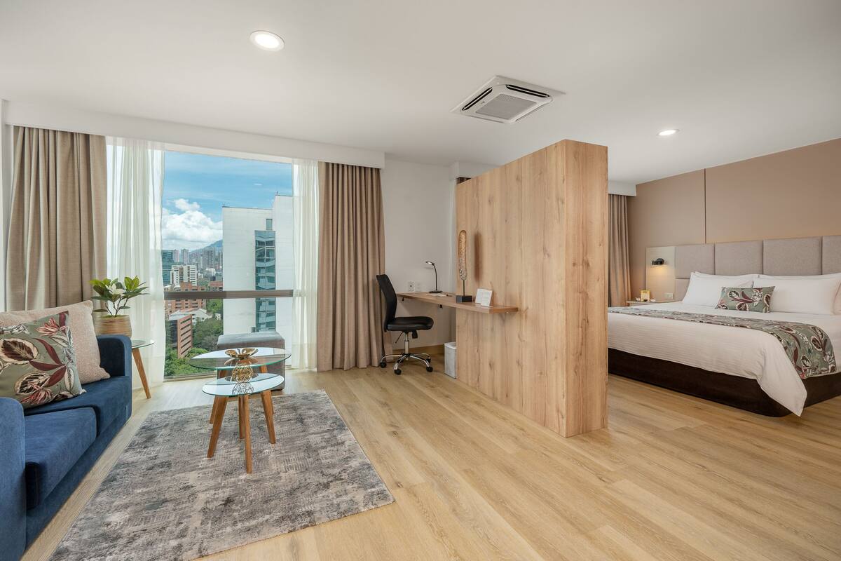 A modern hotel room with a large bed, a desk, a blue sofa, and a view of the city.