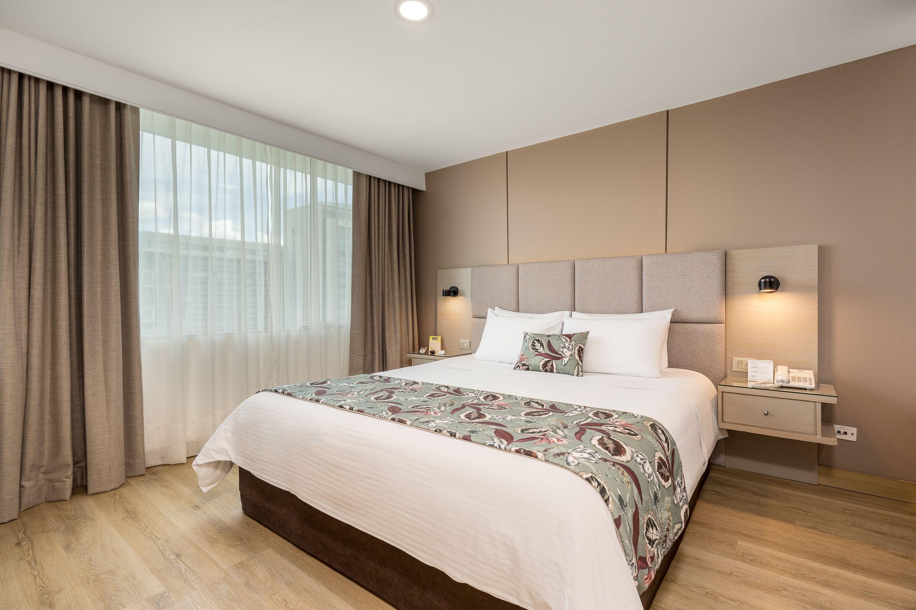 A modern hotel room with a large bed, a nightstand, a wall-mounted lamp, and a view of the city through large windows.