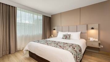 A modern hotel room with a large bed, a nightstand, a wall-mounted lamp, and a view of the city through large windows.