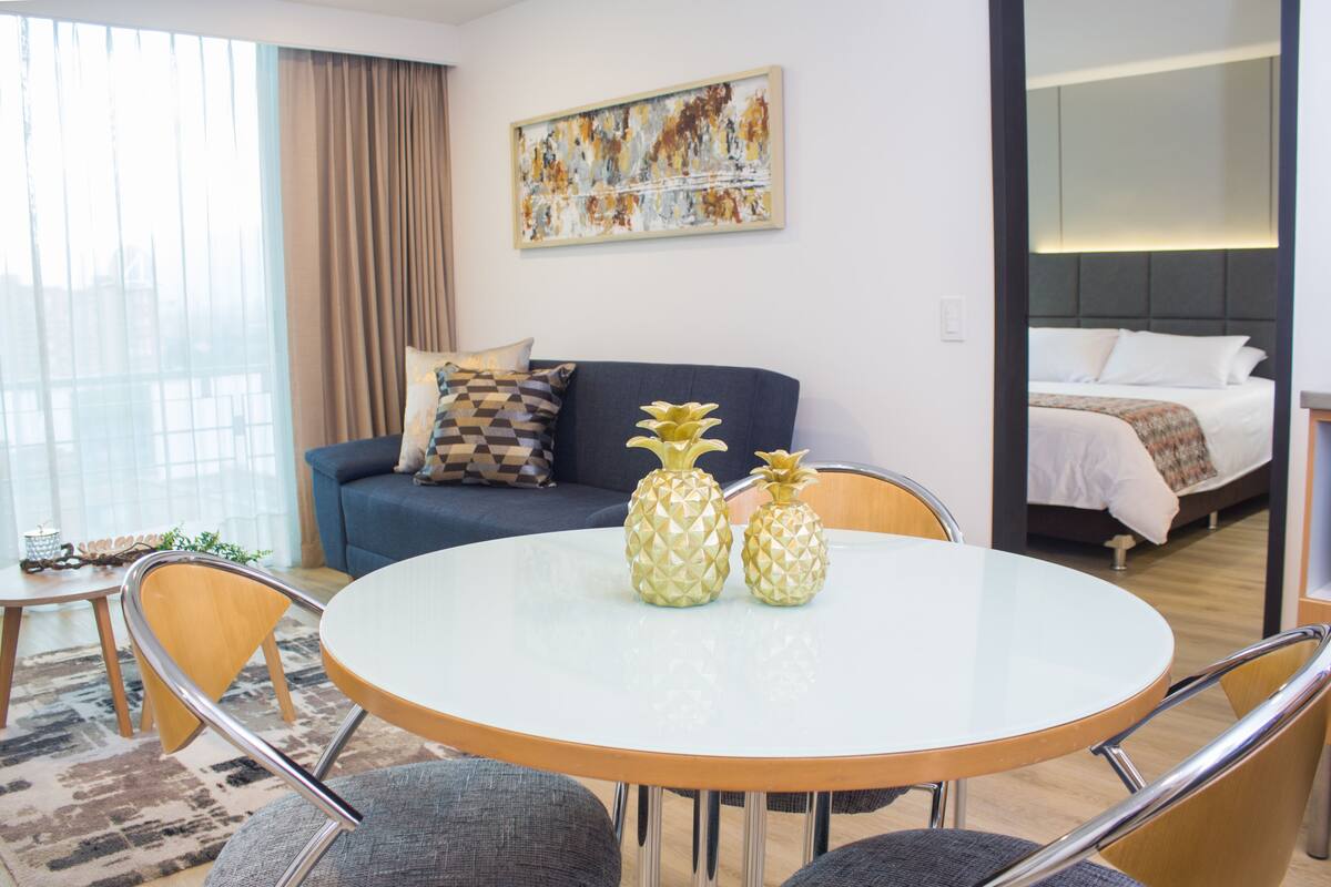 A modern hotel room with a round dining table, two pineapple-shaped vases, a blue sofa with patterned cushions, a large window with curtains, and a painting on the wall.