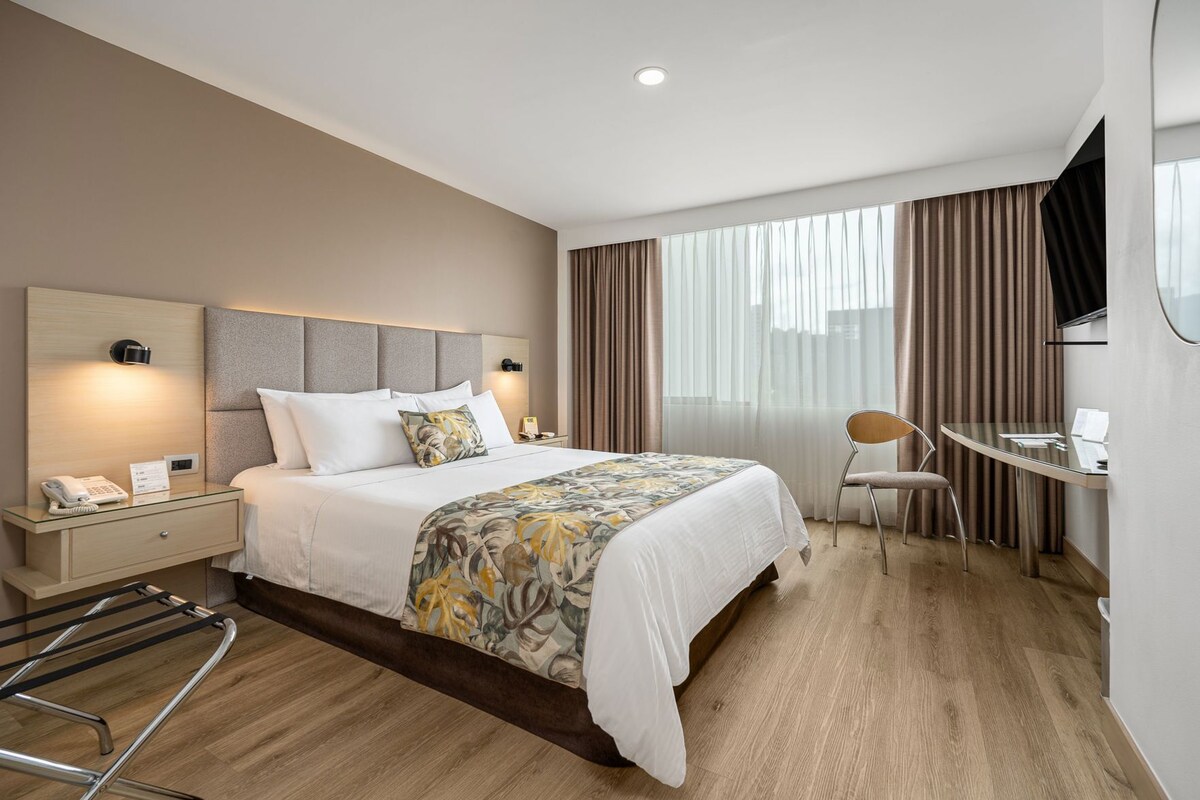 A hotel room with a bed, bedside table, chair, desk, and a TV mounted on the wall.