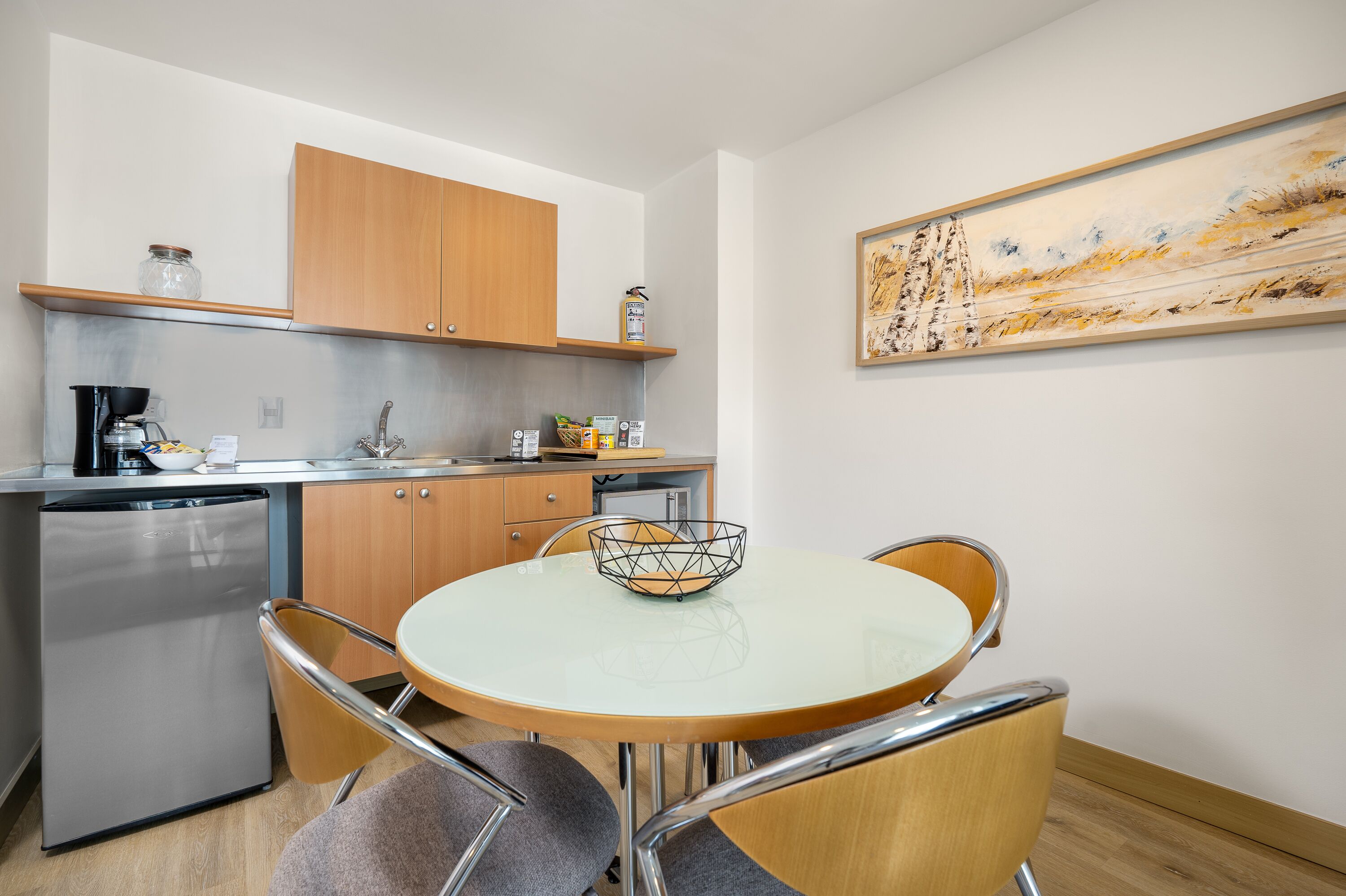 A compact kitchen with a dishwasher, a coffee machine, and a microwave. There is a round table with two chairs and a decorative metal bowl. A painting is on the wall above the table.