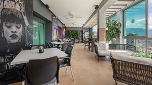 A modern dining area with high tables and chairs, a large mural on the wall, and a view of the city through large windows. - Novelty Suites Hotel - Oviedo Mall (Medellín)