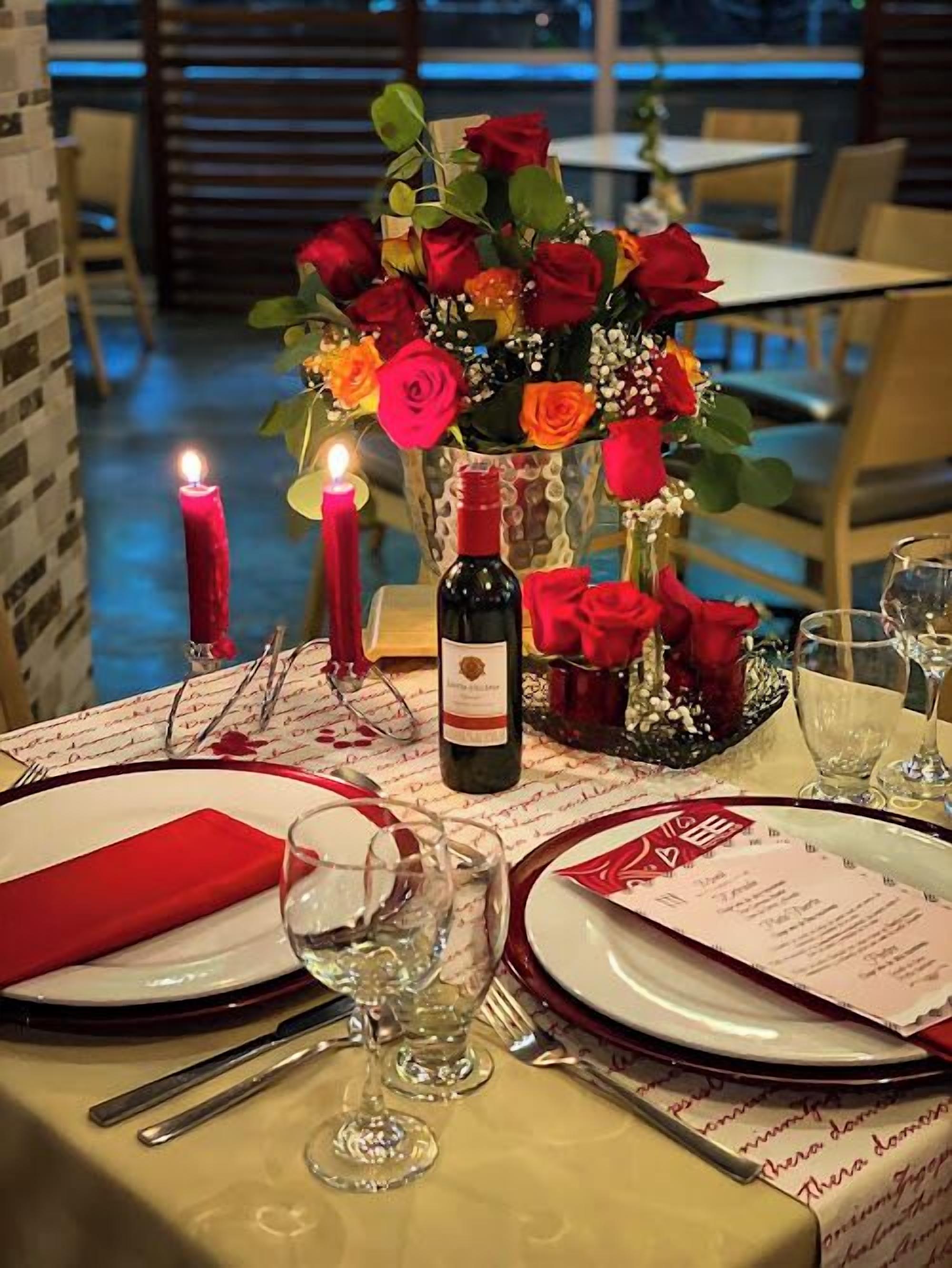 Une table dressée pour deux personnes, avec une bougie, une bouteille de vin et un vase de roses rouges.