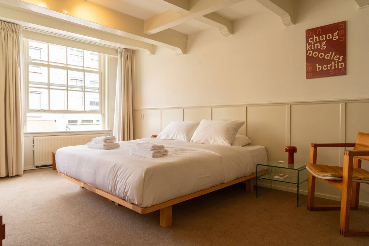A bedroom with a large window, a bed with white linens, a wooden nightstand, and a chair.