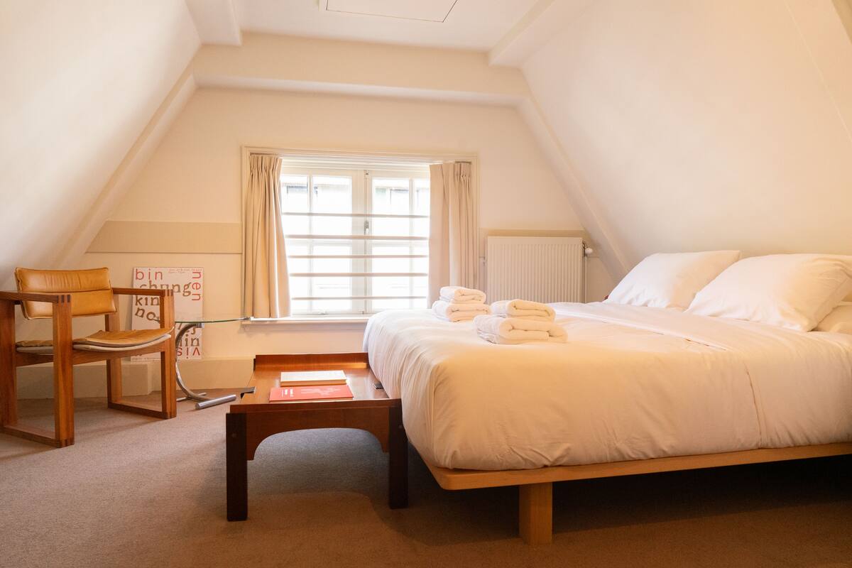 A bedroom with a bed, a chair, a table, and a window with curtains.