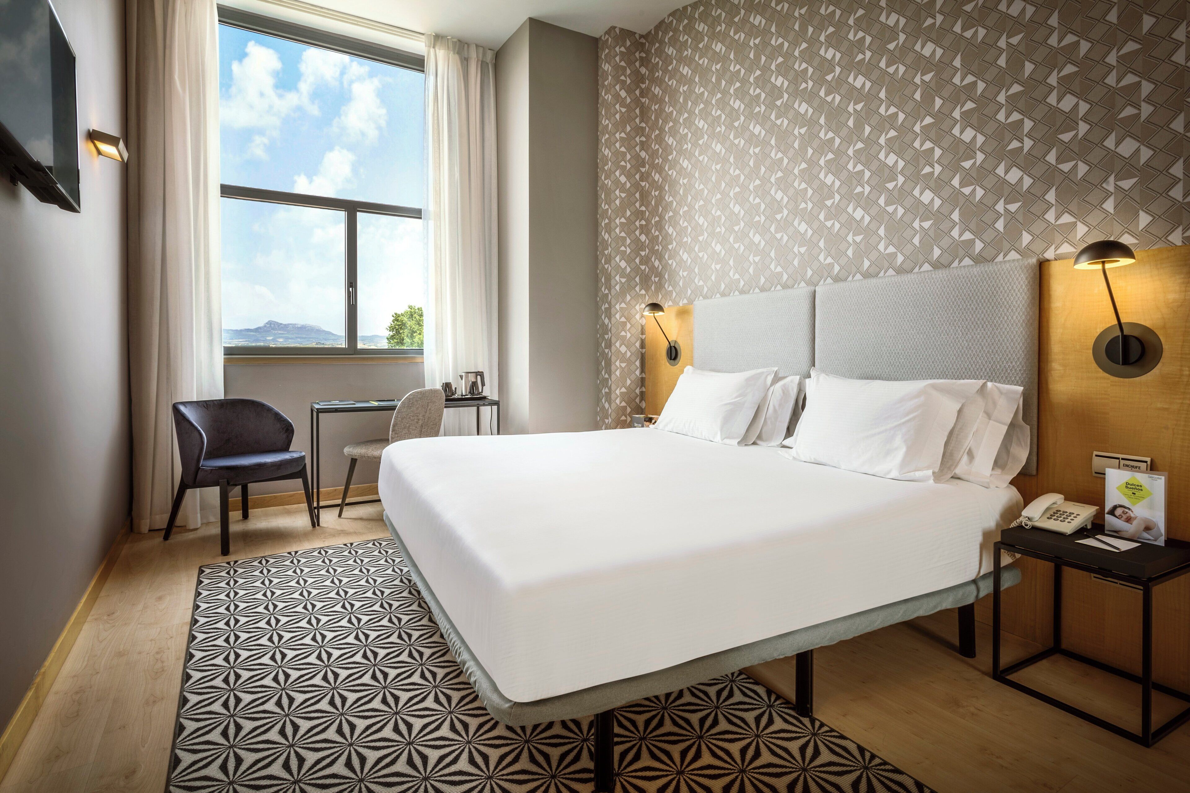 A hotel room with a large bed, a desk with a chair, a window with curtains, and a patterned wall.