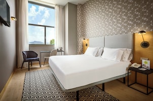 Quarto de hotel com uma cama grande, uma mesa com cadeira, uma janela com cortinas e um papel de parede estampado.