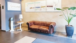 A lobby with a brown leather sofa, a large potted plant, a wall-mounted sign reading "Sercotel", and a framed map. - Hotel Sercotel Portales (Logroño)