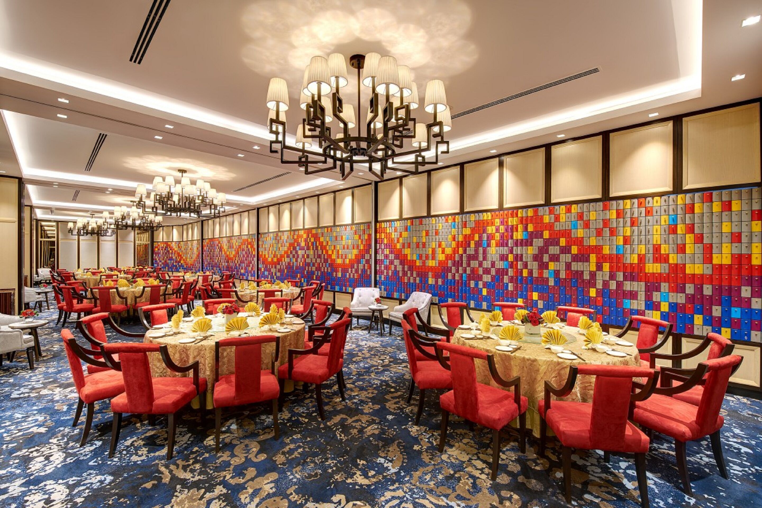 A hotel dining area with round tables, red chairs, and a colorful wall art installation.