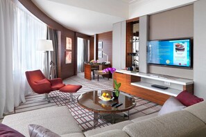 Una habitación de hotel moderna con un sofá, un sillón y una mesa de centro redonda con fruta. Un televisor de pantalla plana está montado en la pared, y hay un pequeño escritorio con una lámpara.