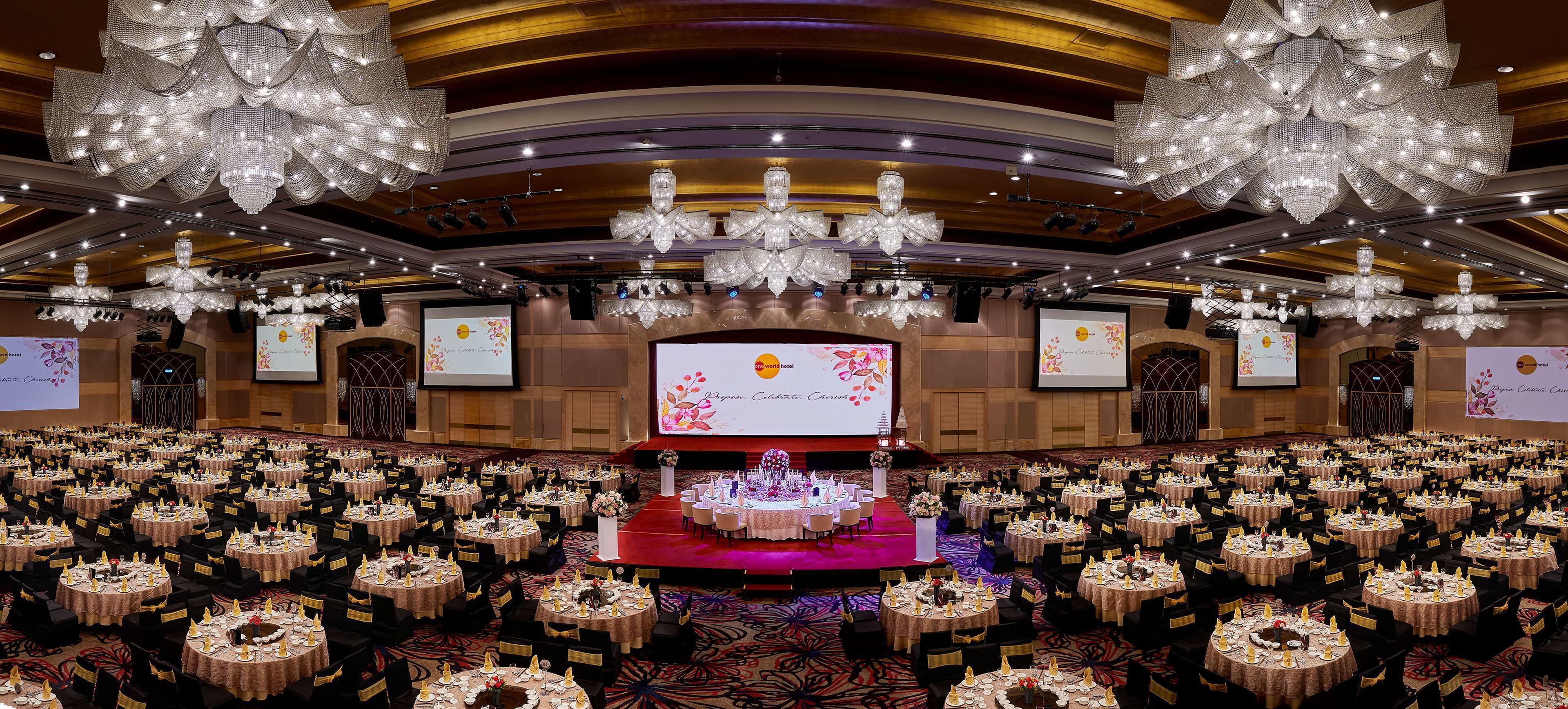A large banquet hall with round tables, chandeliers, and a stage with a screen.