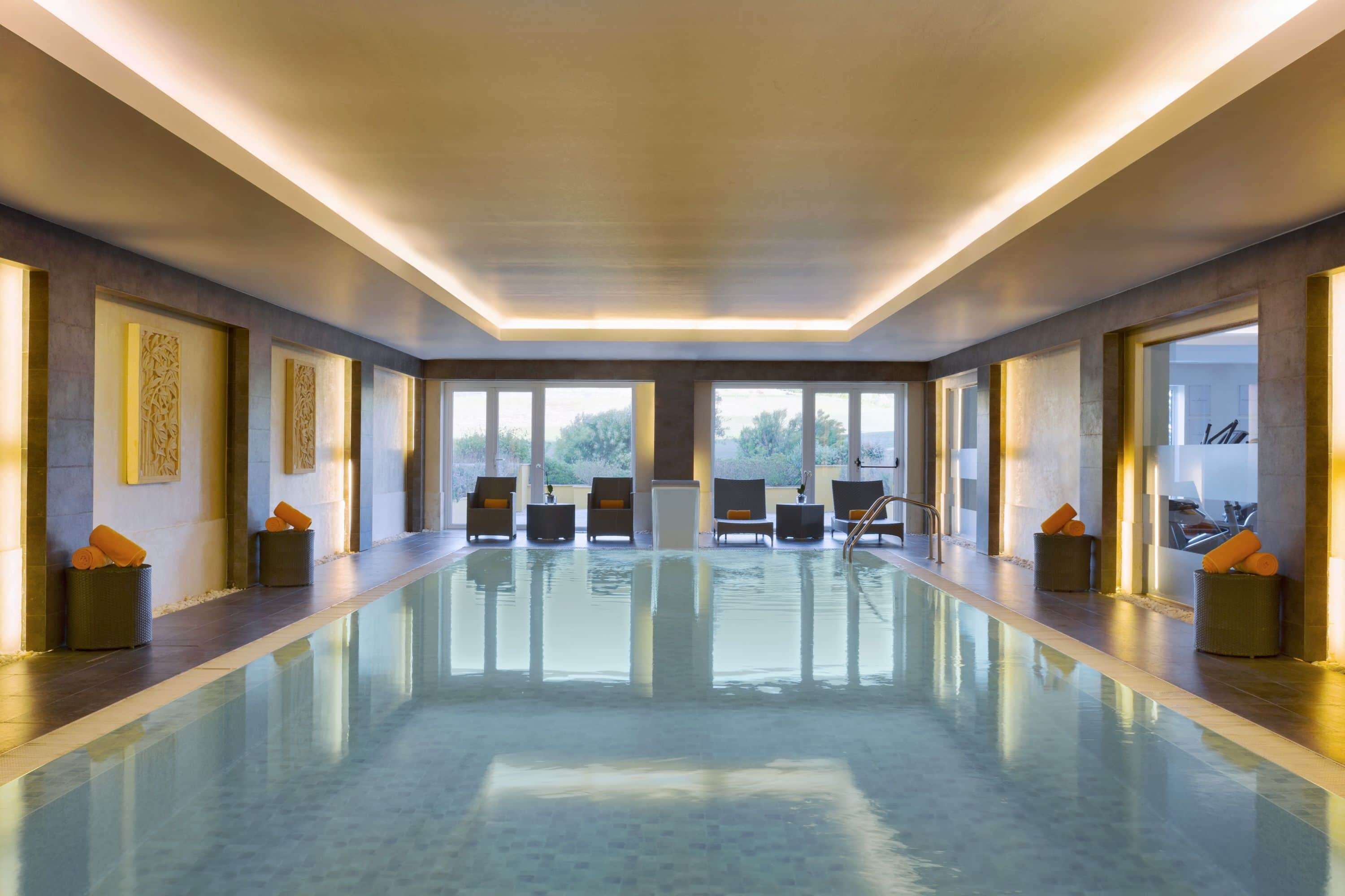 Una larga piscina interior de diseño moderno, con un pasillo central y zonas de estar con cojines naranja.
