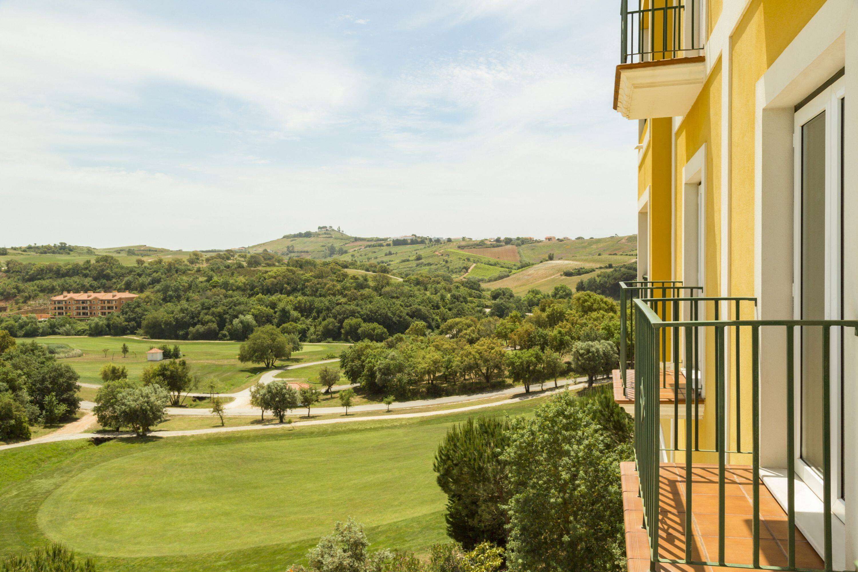 A yellow building with a balcony overlooking a green landscape with trees and hills.