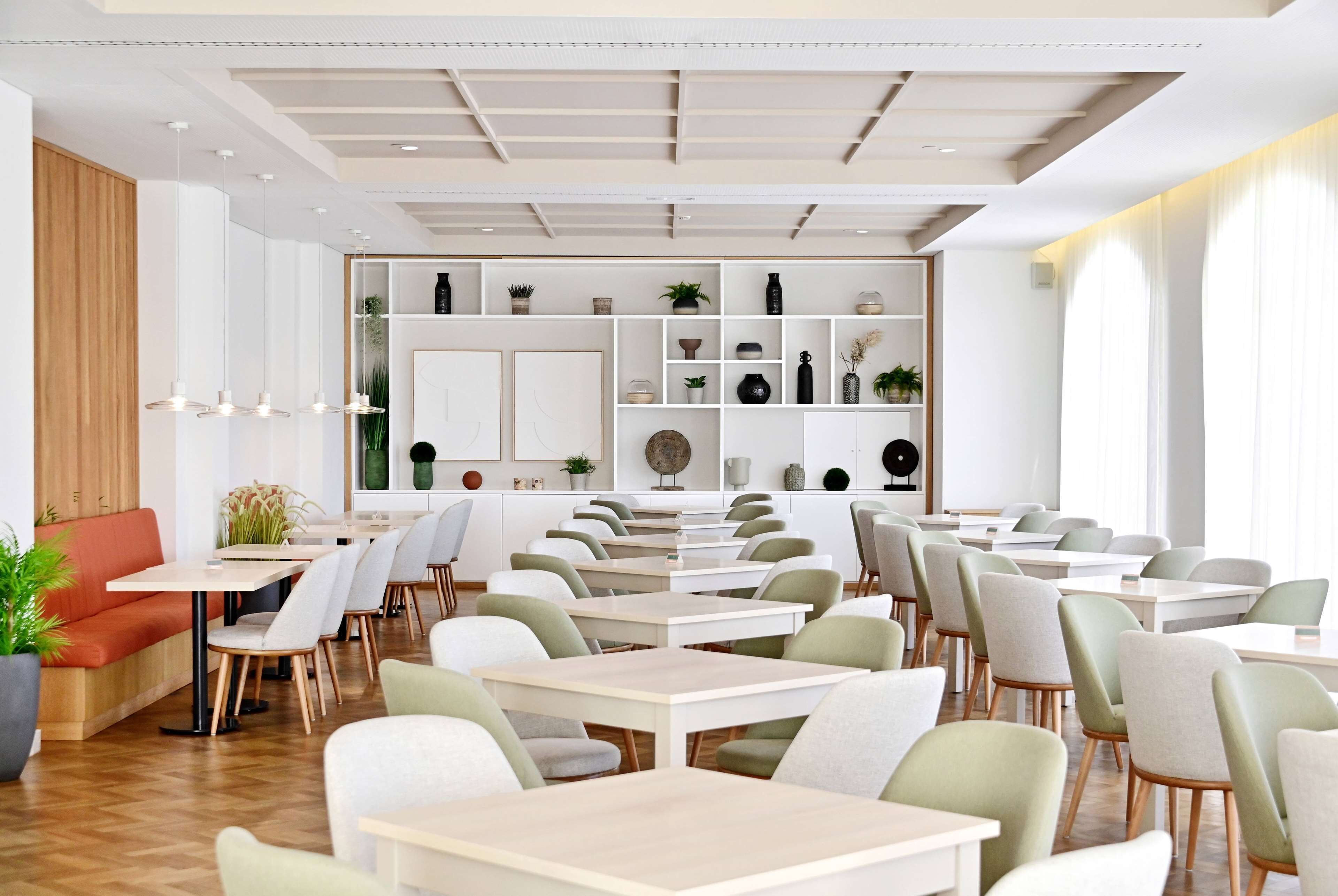 A modern restaurant with a clean, minimalist design, featuring white and light green furniture, a variety of plants on shelves, and a bright, well-lit interior.