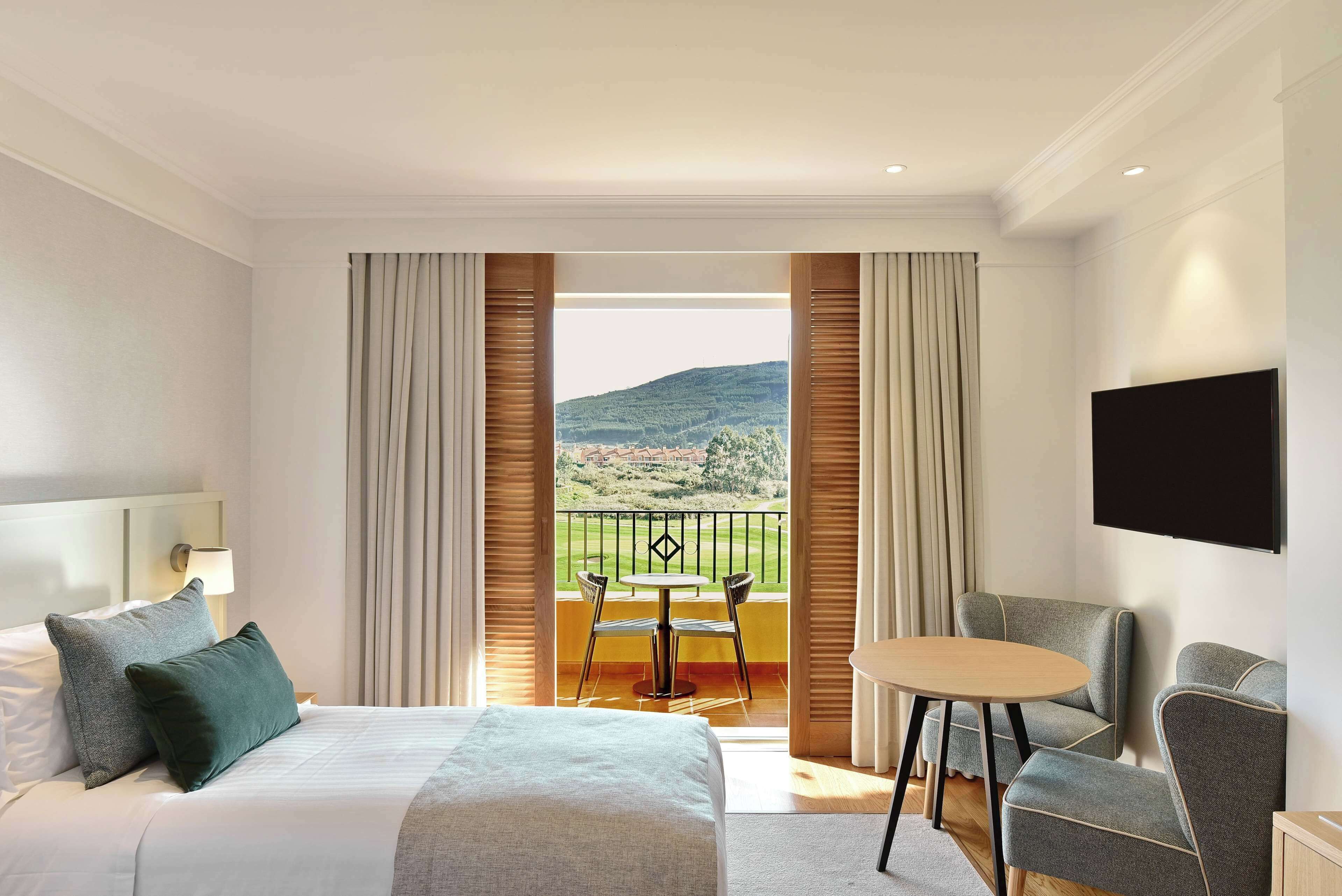 Une chambre d’hôtel équipée d’un lit, d’un bureau, d’une chaise, d’une télévision et d’un balcon offrant une vue sur la campagne.