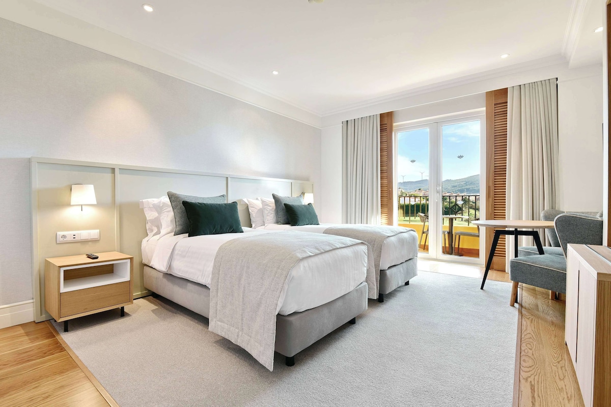 A modern hotel room with two beds, a desk, and a balcony with a view.