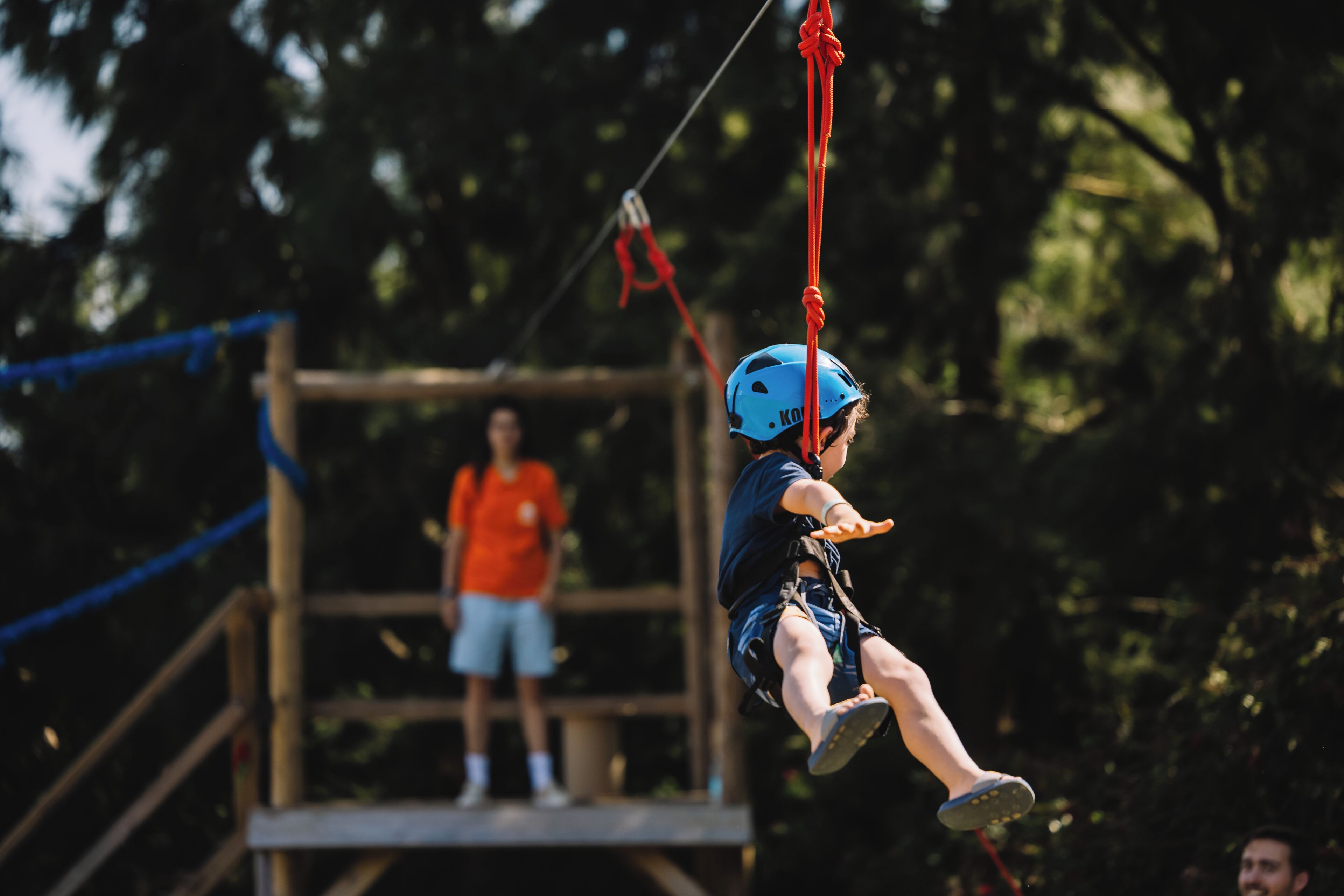 A person on a zip line, wearing a blue helmet, with a safety harness and a harness attached to a red rope.