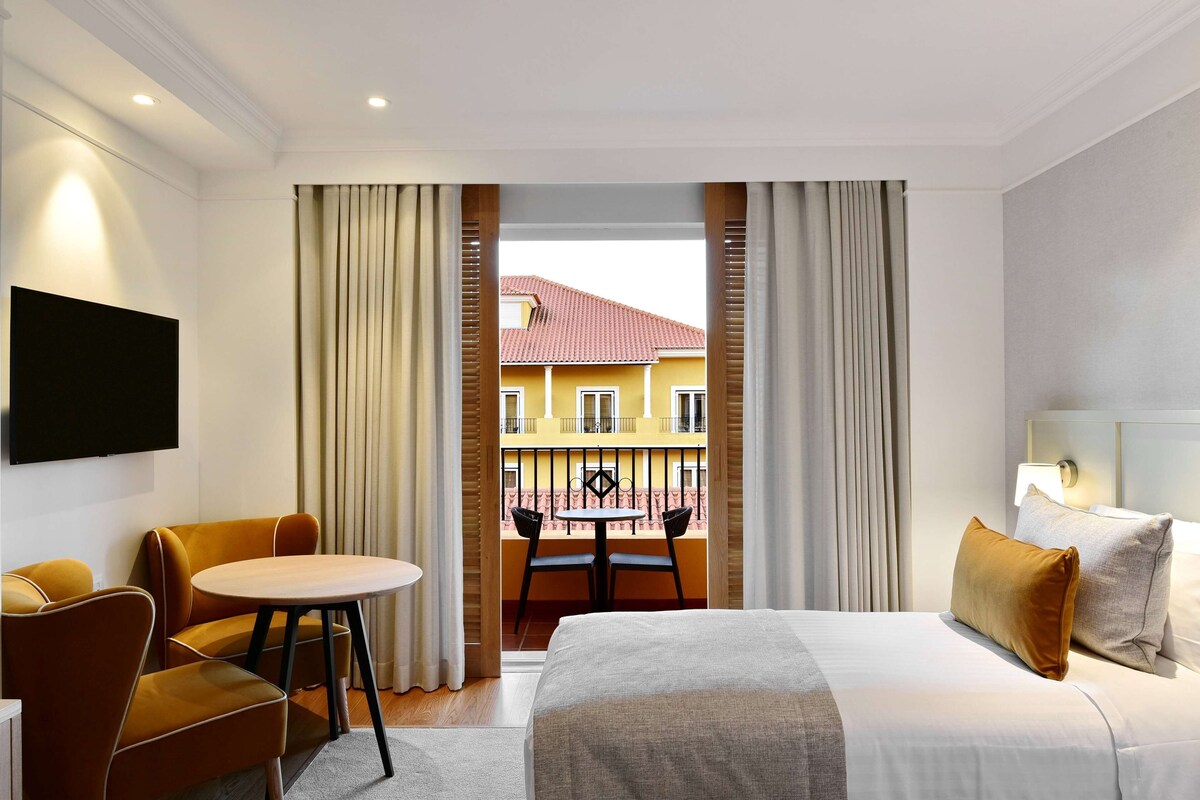 A hotel room with a bed, a round table with chairs, a TV mounted on the wall, and a balcony with a view of a yellow building.