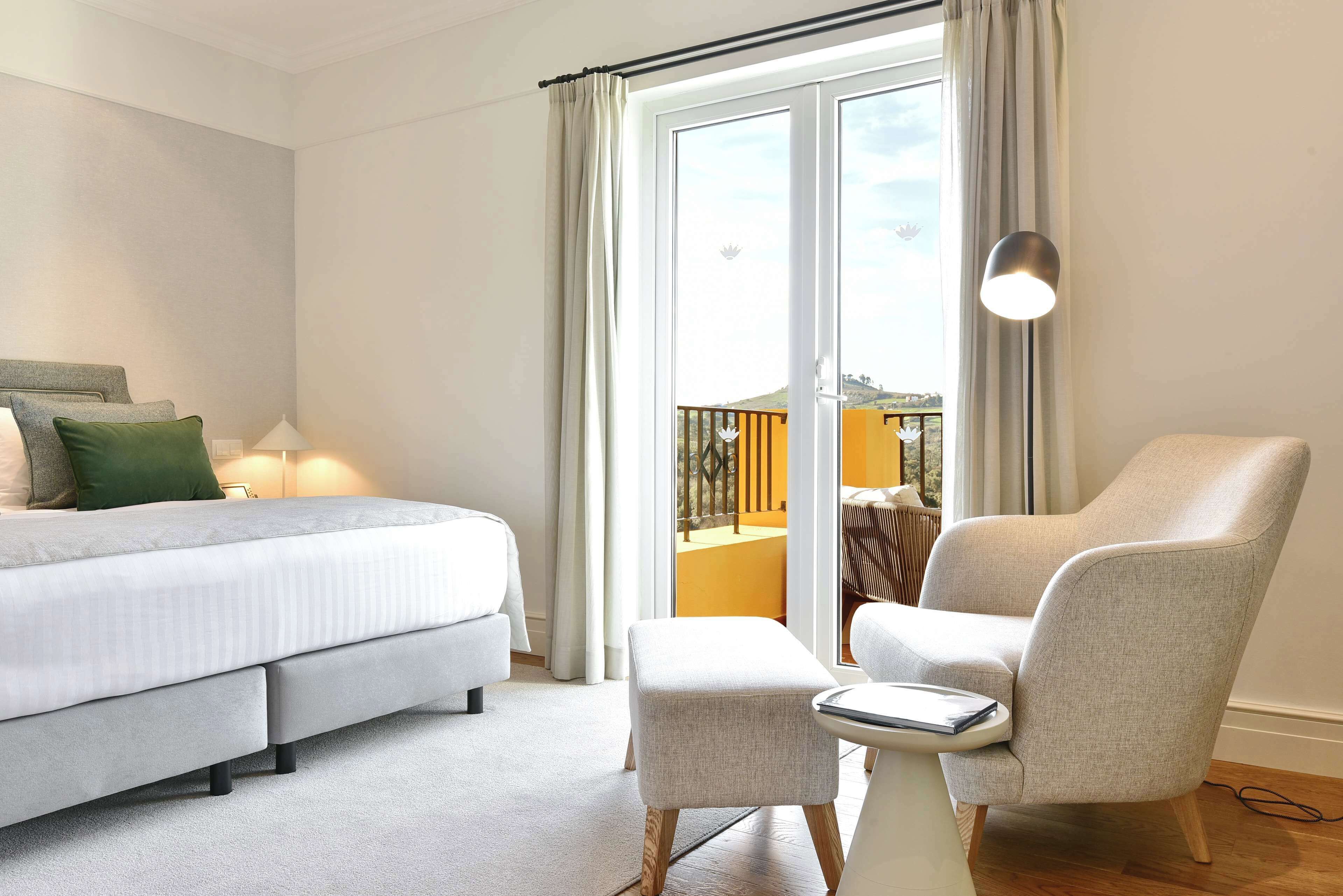 A modern hotel room with a bed, a small table, a chair, and a balcony with a view.