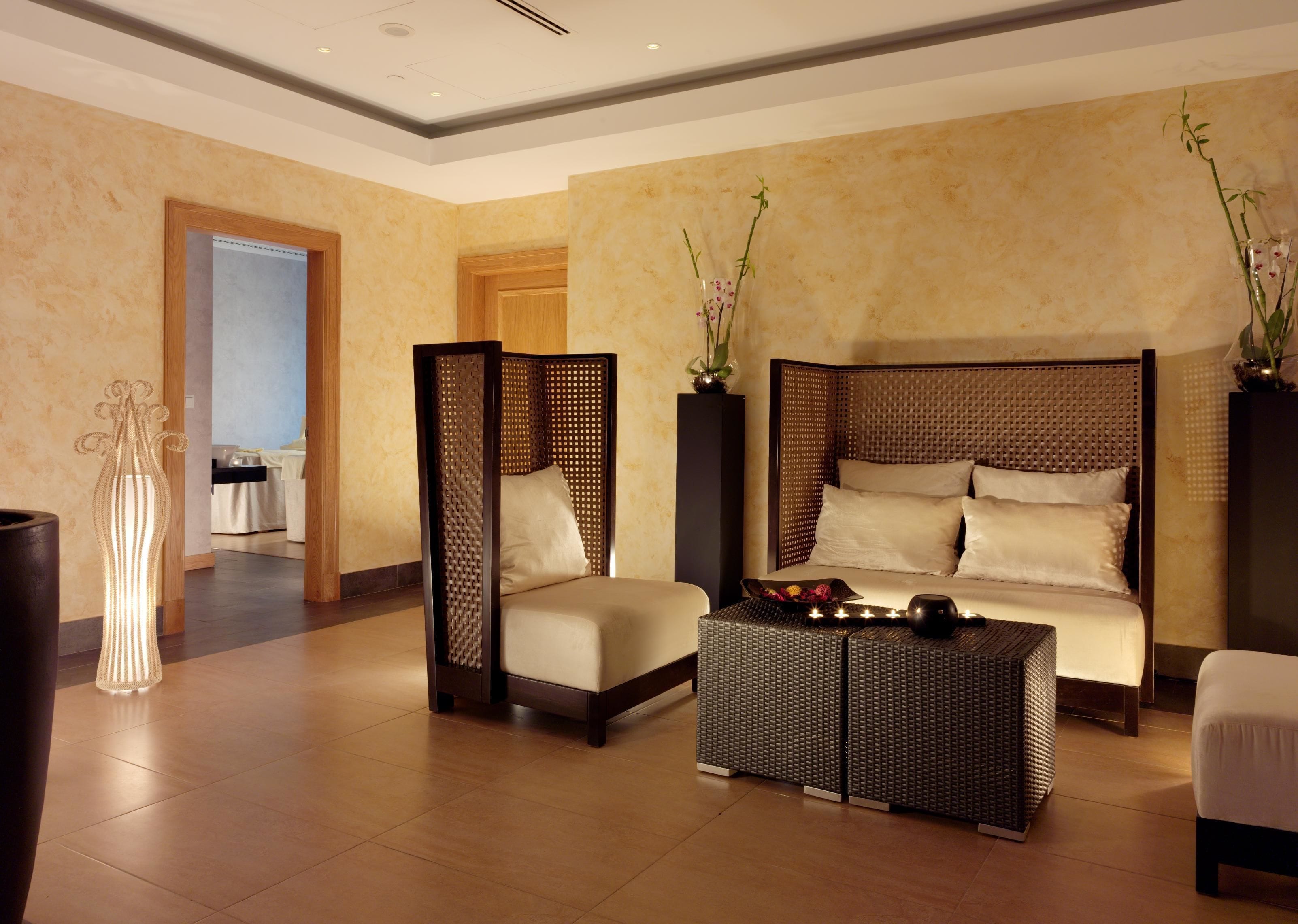 A modern hotel room with a large bed, a sitting area with a sofa and armchairs, a decorative floor lamp, and a view of the adjacent room.