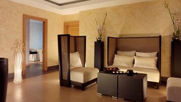 A modern hotel room with a large bed, a sitting area with a sofa and armchairs, a decorative floor lamp, and a view of the adjacent room.