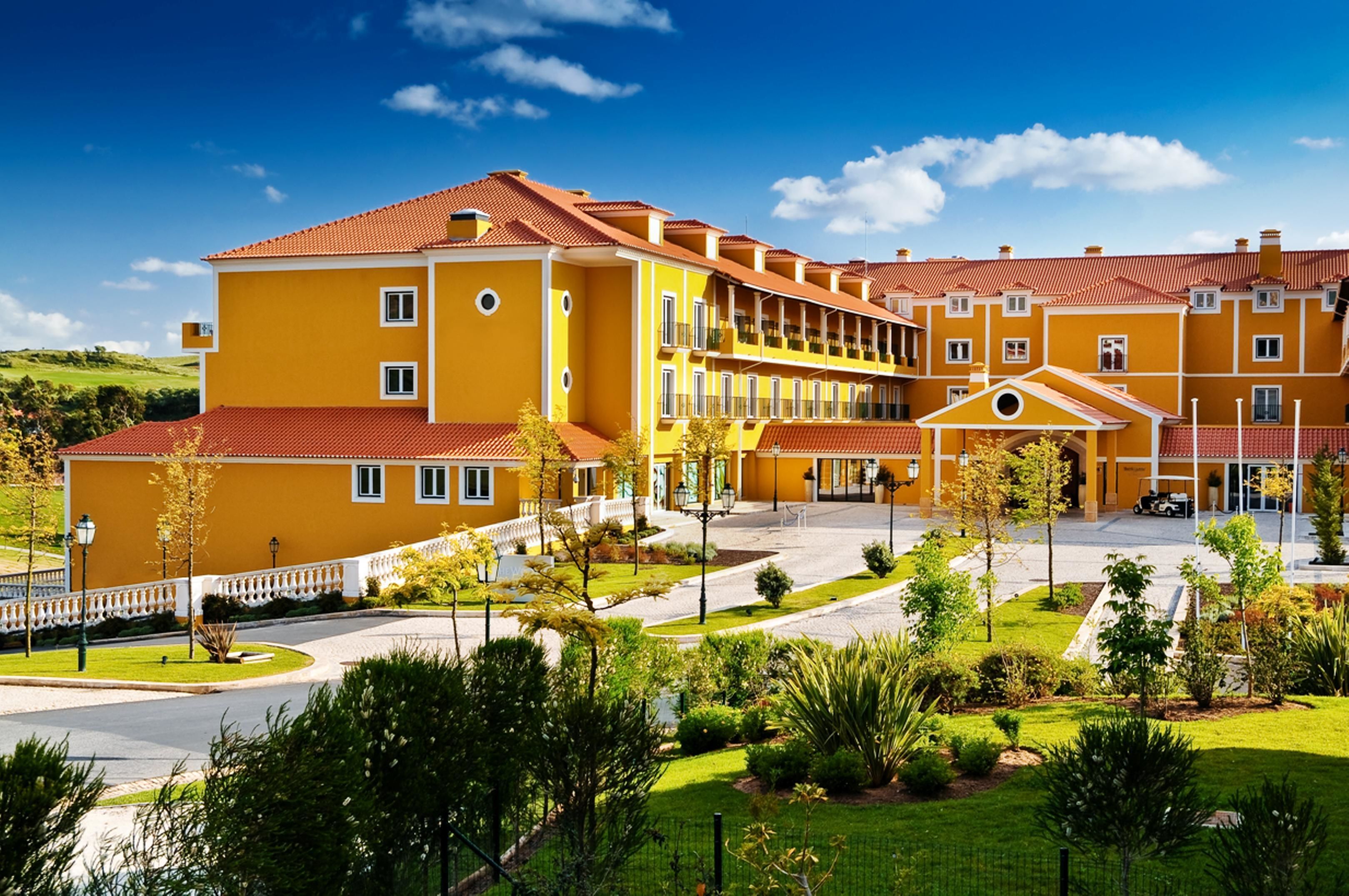 A yellow building with multiple balconies and a red roof, surrounded by green landscaping and a clear blue sky.