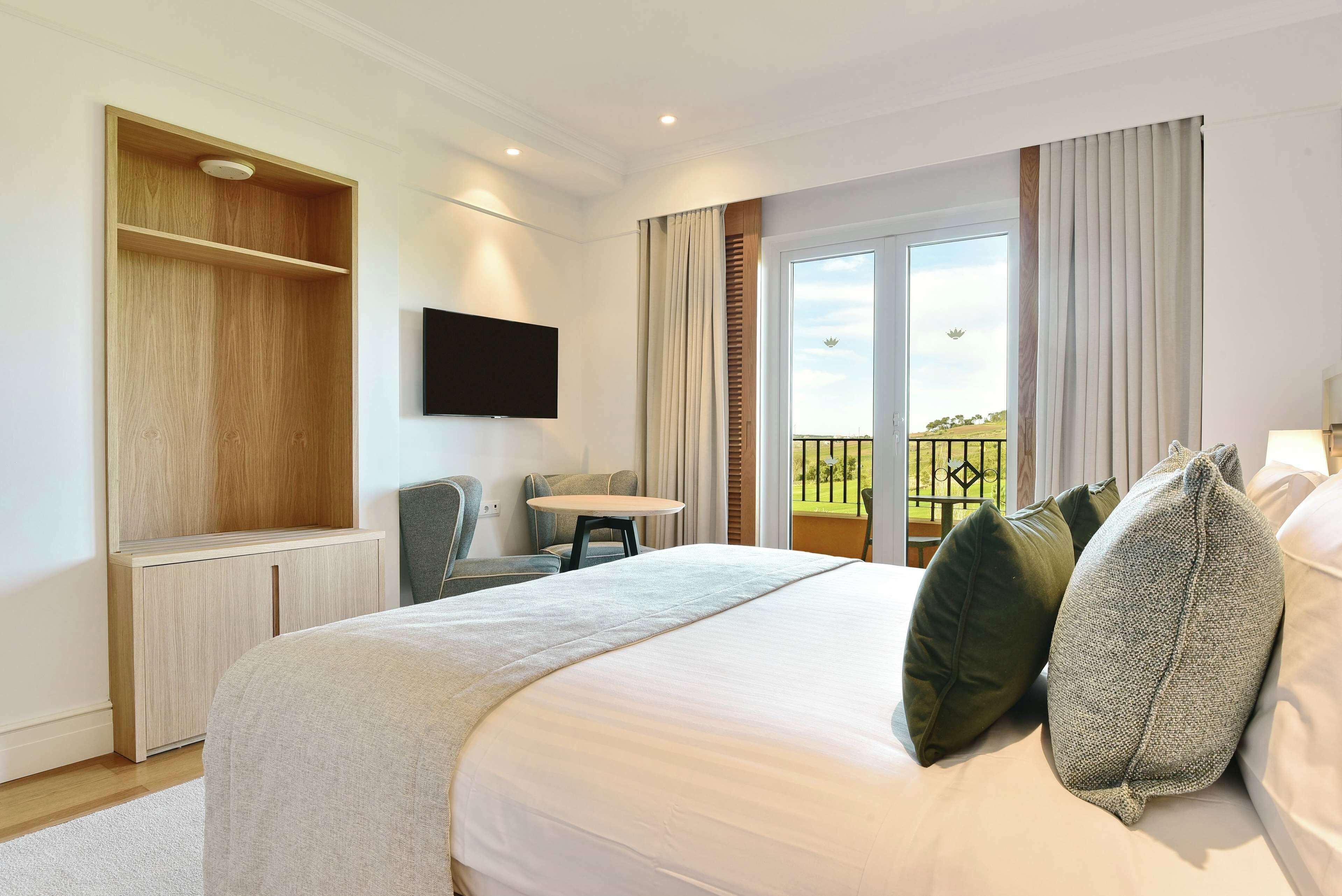 A hotel room with a large bed, a desk, a chair, a TV mounted on the wall, and a balcony with a view.