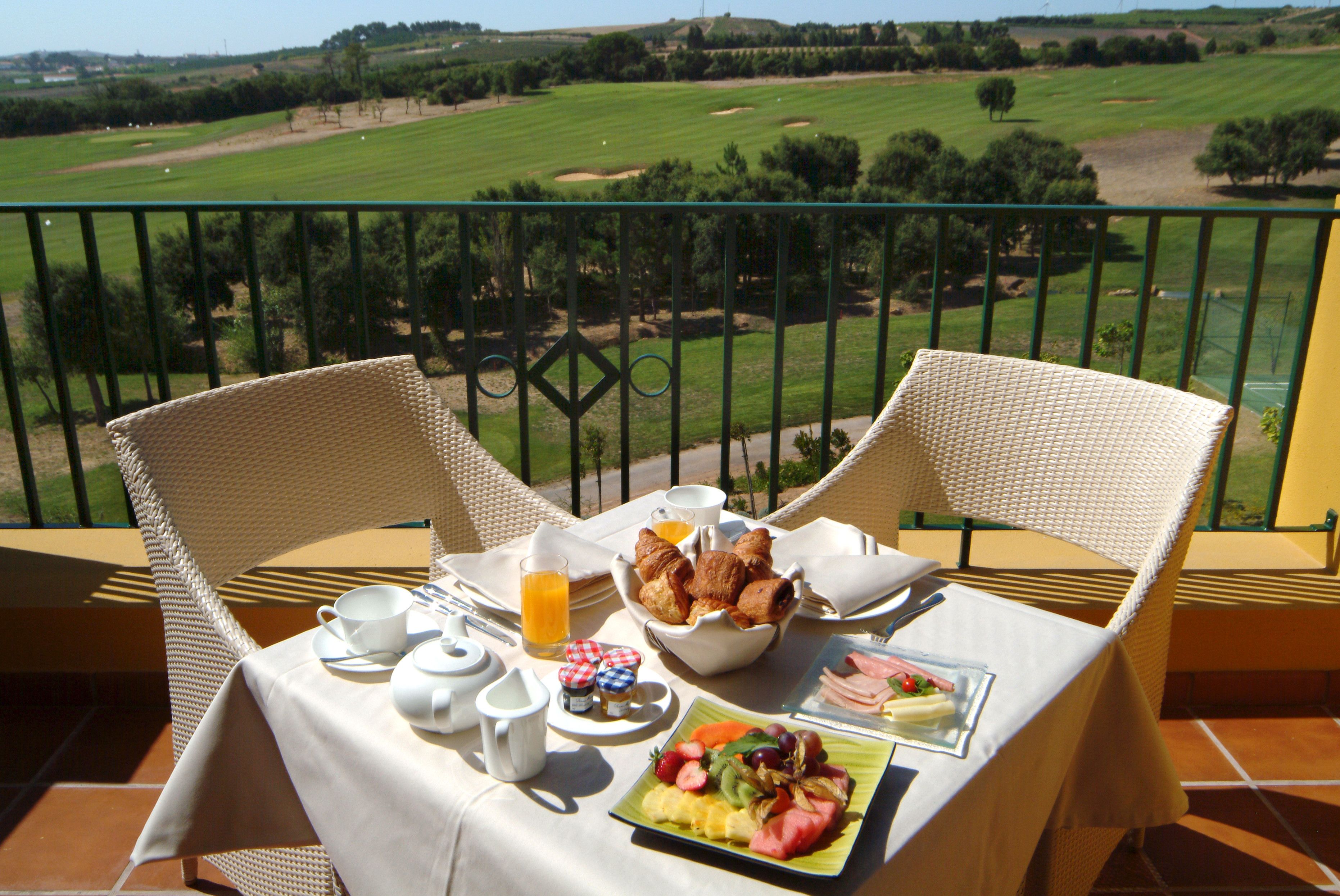 Mesa en el balcón con artículos para el desayuno, incluyendo cruasanes, fruta y un plato de jamón y huevos, con vistas a un campo de golf.