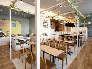 Un café moderno con mesas y sillas de madera, una zona de cocina y plantas colgando del techo.