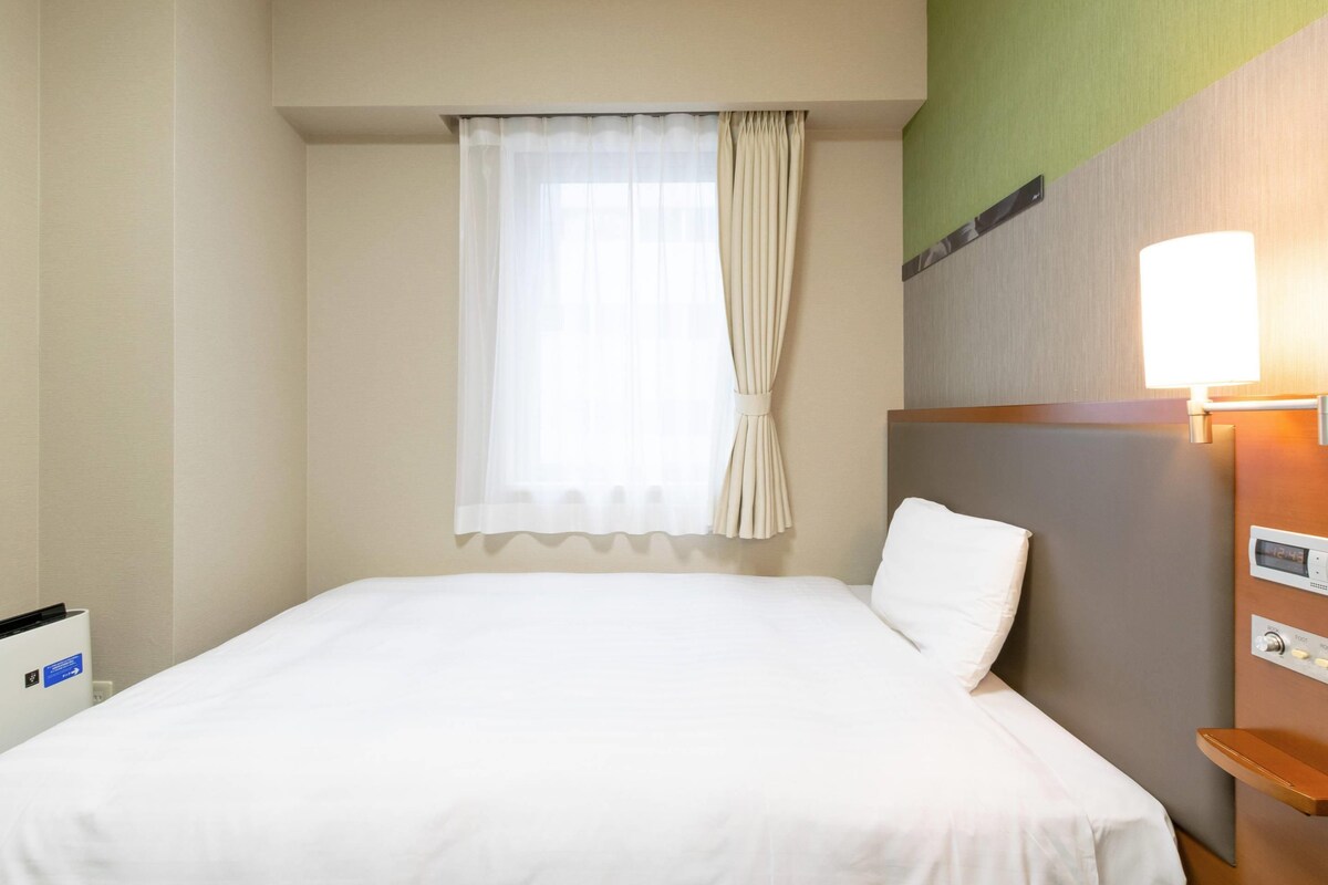 A hotel room with a bed, a window with curtains, a bedside table with a lamp, and a wall-mounted control panel.