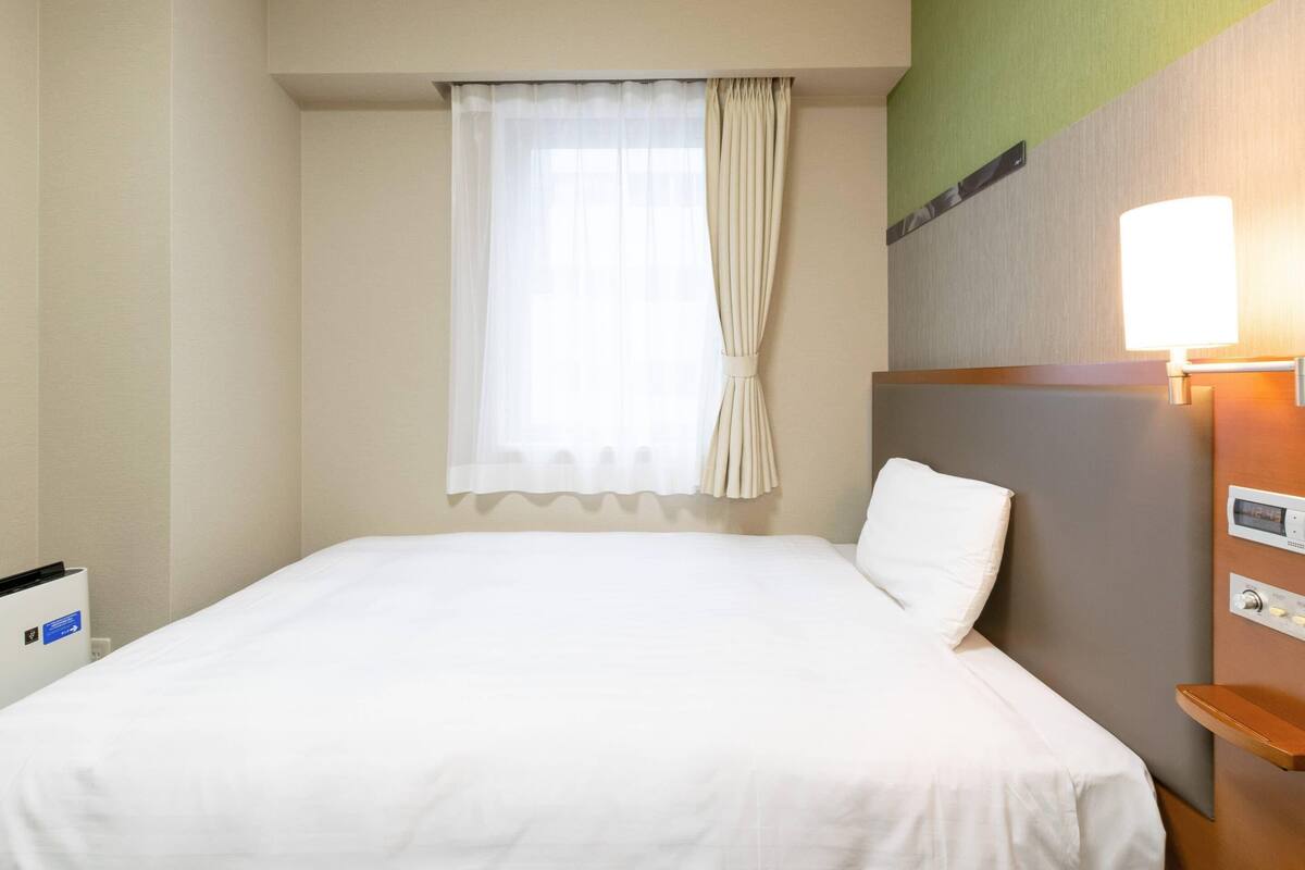 A hotel room with a bed, a window with curtains, a bedside table, and a wall-mounted control panel.