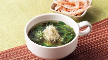 A bowl of soup with green vegetables and a white cup on a textured surface.