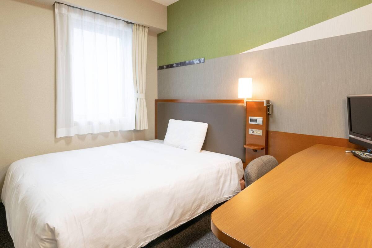 A hotel room with a single bed, a desk with a chair, a television, and a wall-mounted light.