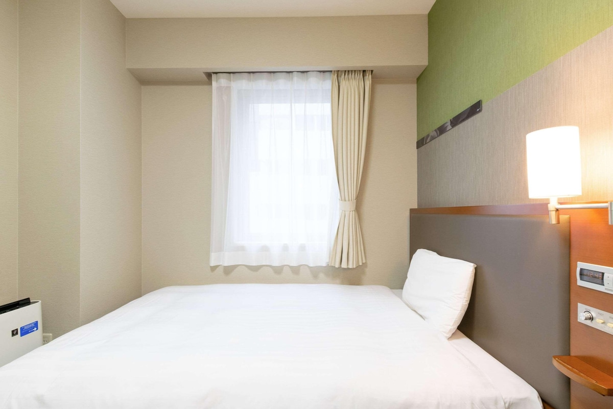 A hotel room with a bed, a window with curtains, a wall-mounted lamp, and a control panel.