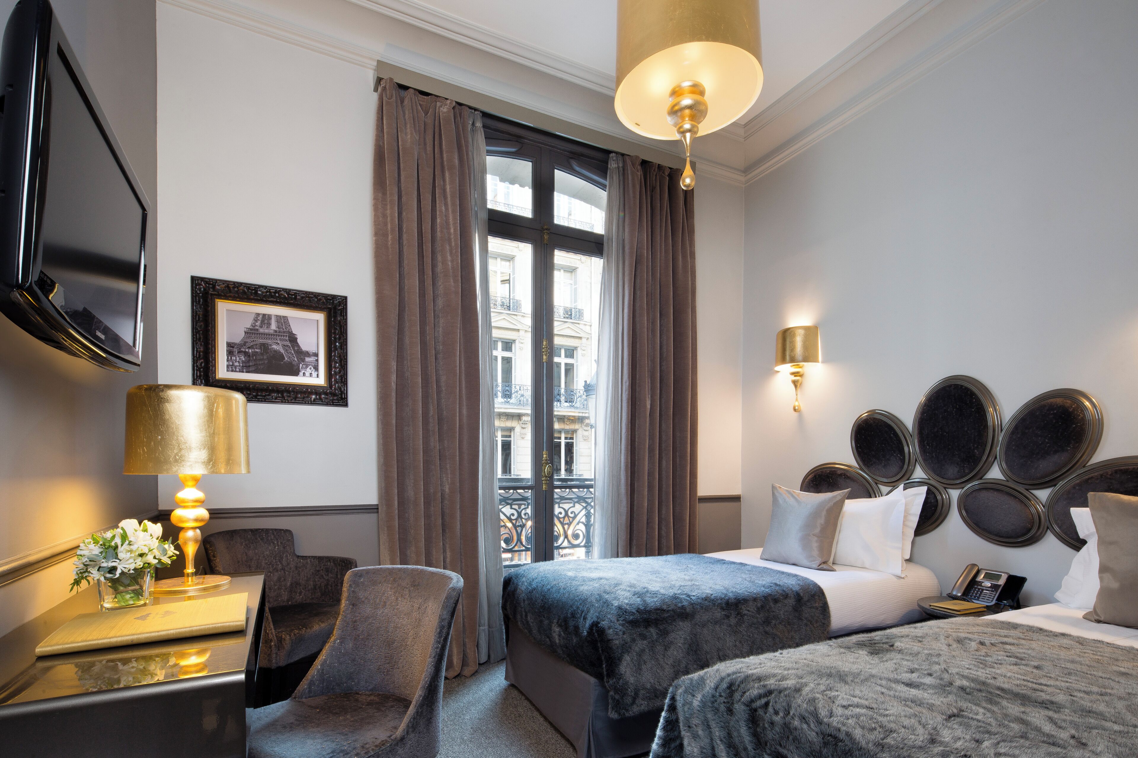 Une chambre d’hôtel avec un lit, un bureau avec une lampe, une chaise, une télévision et une fenêtre avec des rideaux.