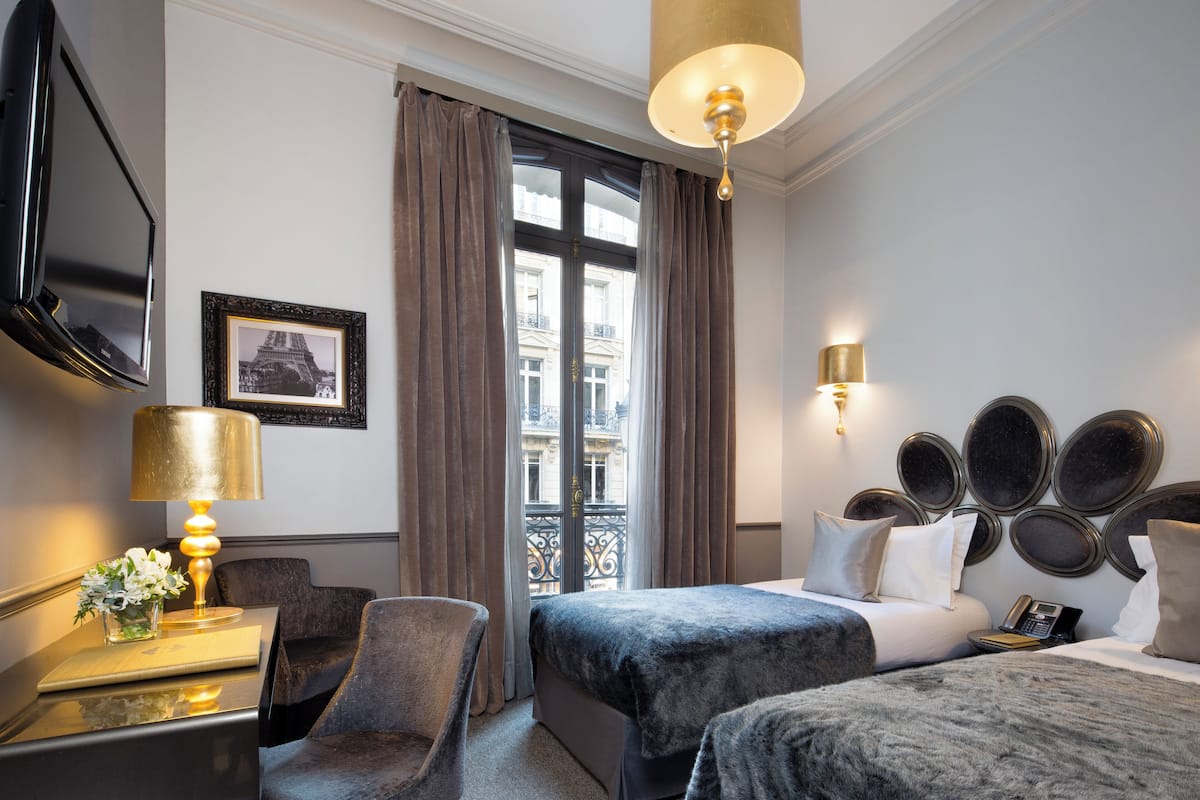 Une chambre d’hôtel avec un lit, un bureau avec une lampe, une chaise, une télévision et une fenêtre avec des rideaux.
