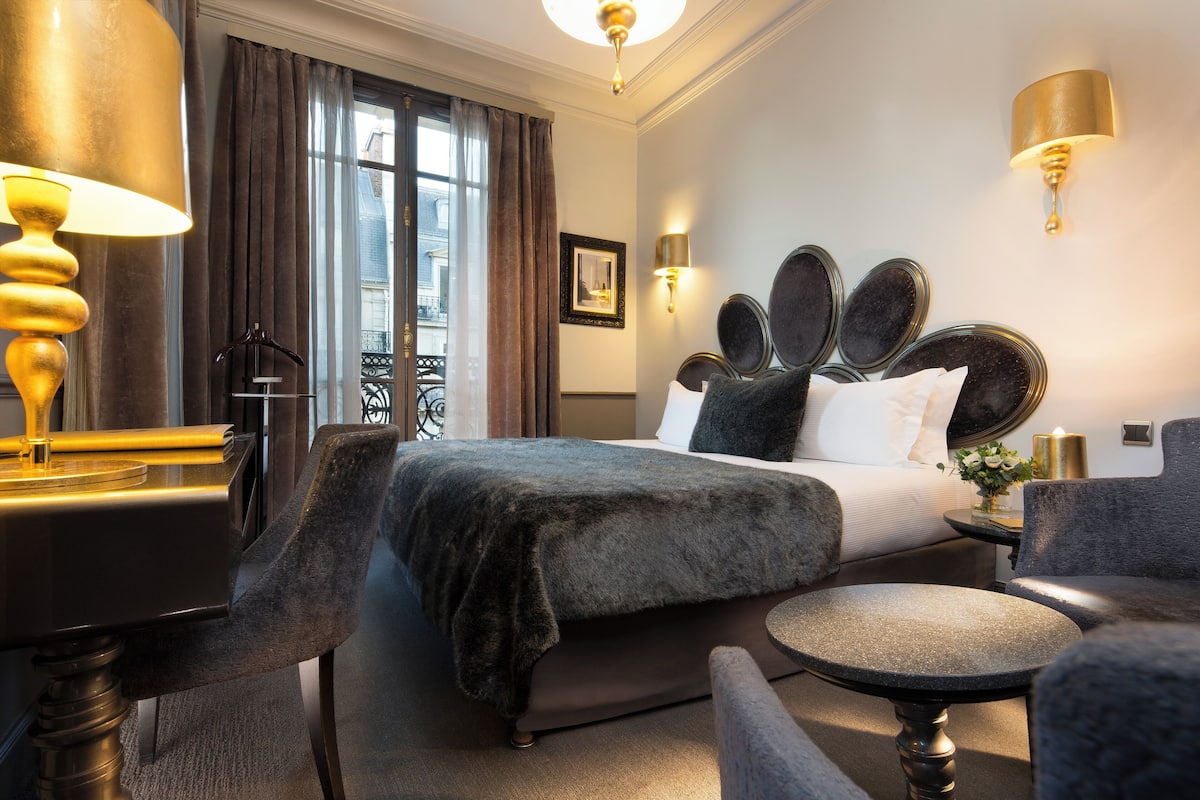 A hotel room with a large bed, a desk with a lamp, a chair, and a small table.