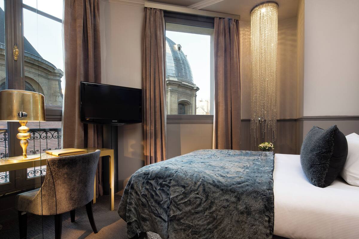 A hotel room with a bed, a desk with a lamp, a television, and a view of a building through the window.
