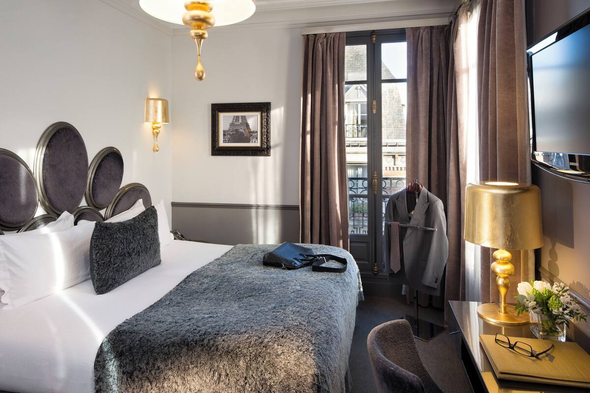 A hotel room with a neatly made bed, a desk with a lamp, a TV, and a view of the city through the window.