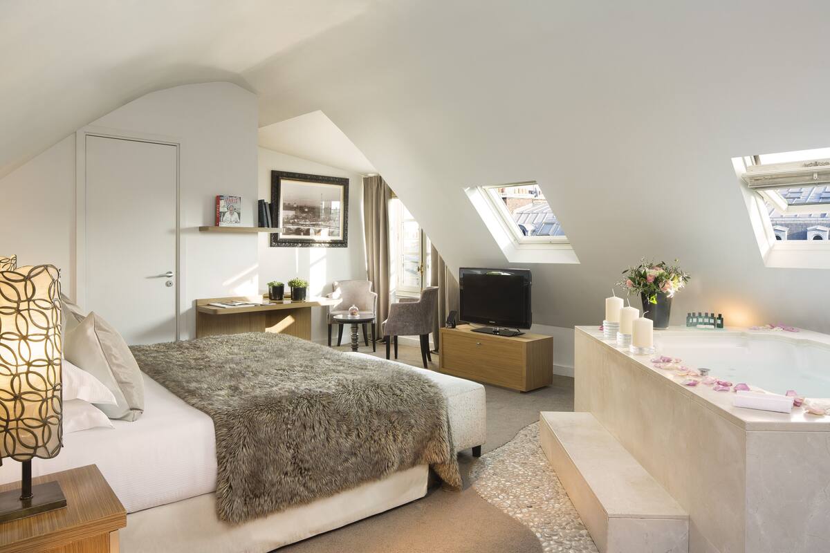 A bedroom with a large bed, a desk, a TV, a bathroom with a bathtub, and skylights.
