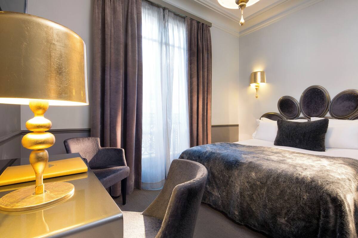 A hotel room with a bed, a desk with a lamp, a chair, and a large window with curtains.