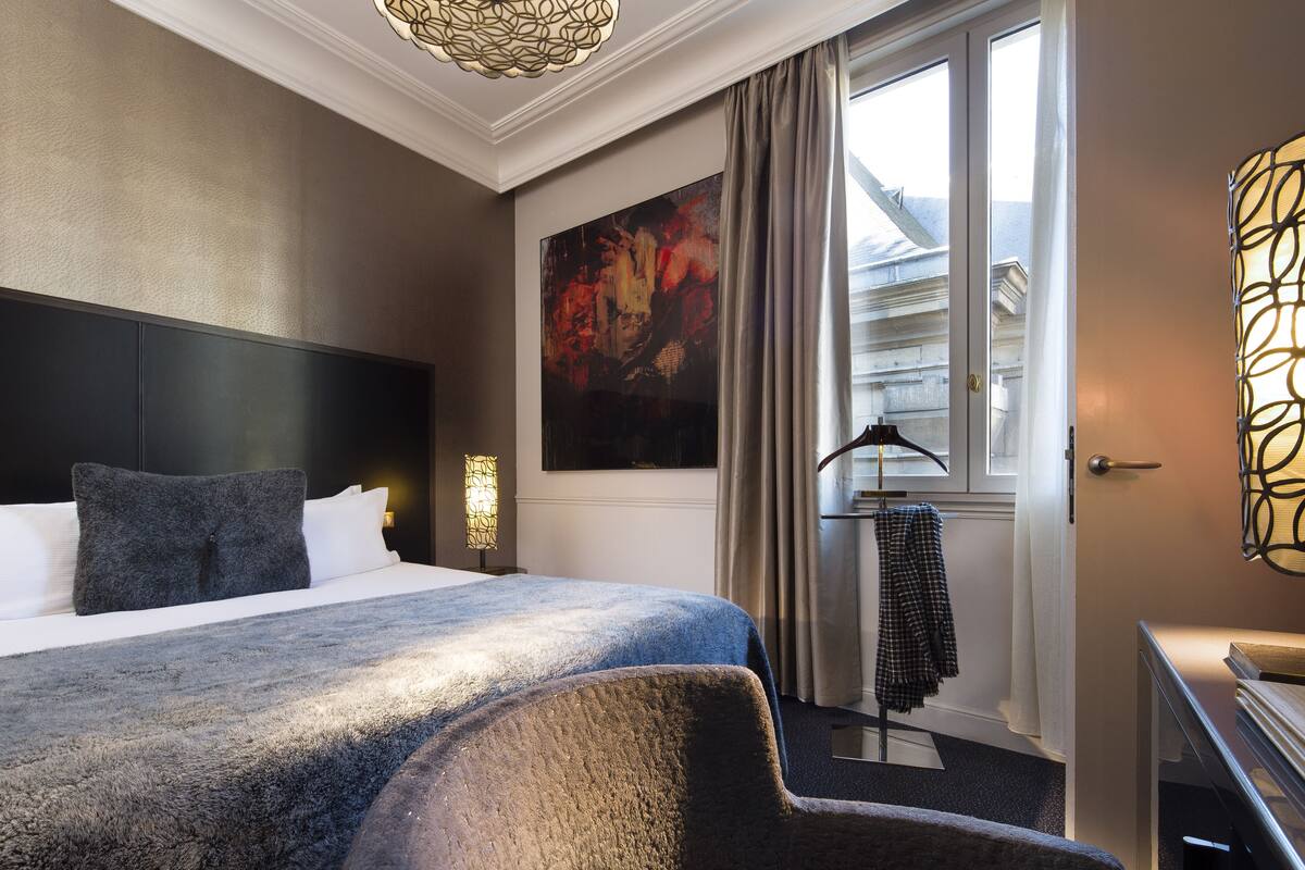 A hotel room with a large bed, a headboard, a painting on the wall, a coat rack, and a desk with a lamp.
