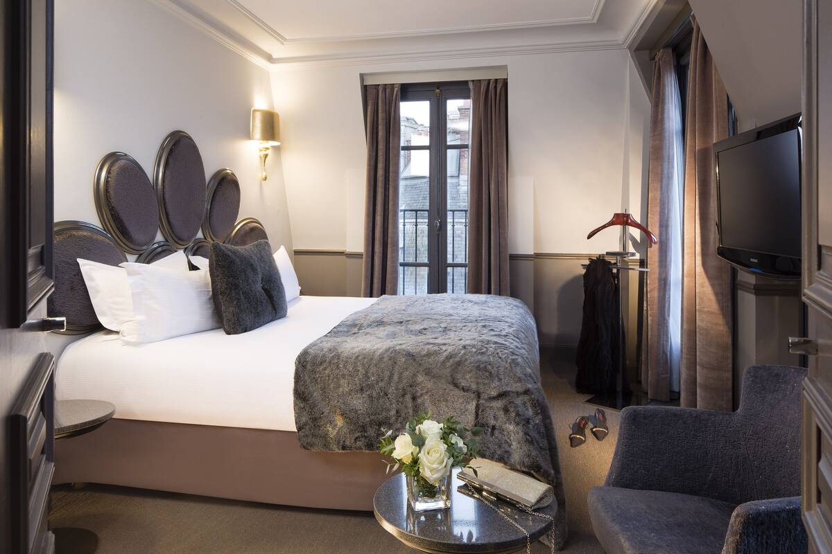 A hotel room with a large bed, a bedside table with a vase of flowers, a chair, a television, and a window with curtains.